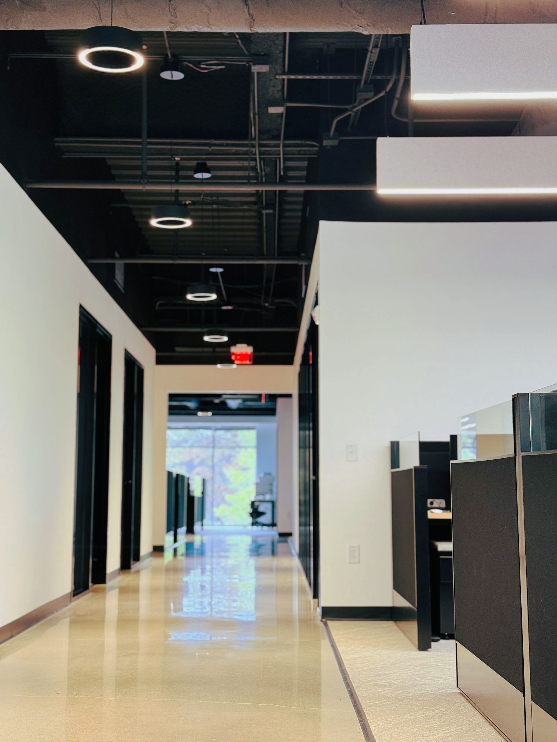 Hallway with white walls, black doors, and exposed ceiling, leading towards an open area with sunlight.