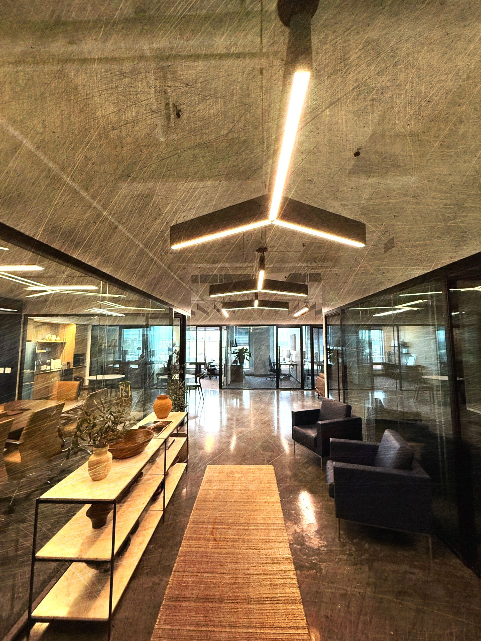 Interior hallway with concrete ceiling, linear lights, glass walls, and a rug runner. Seating and shelving are visible.