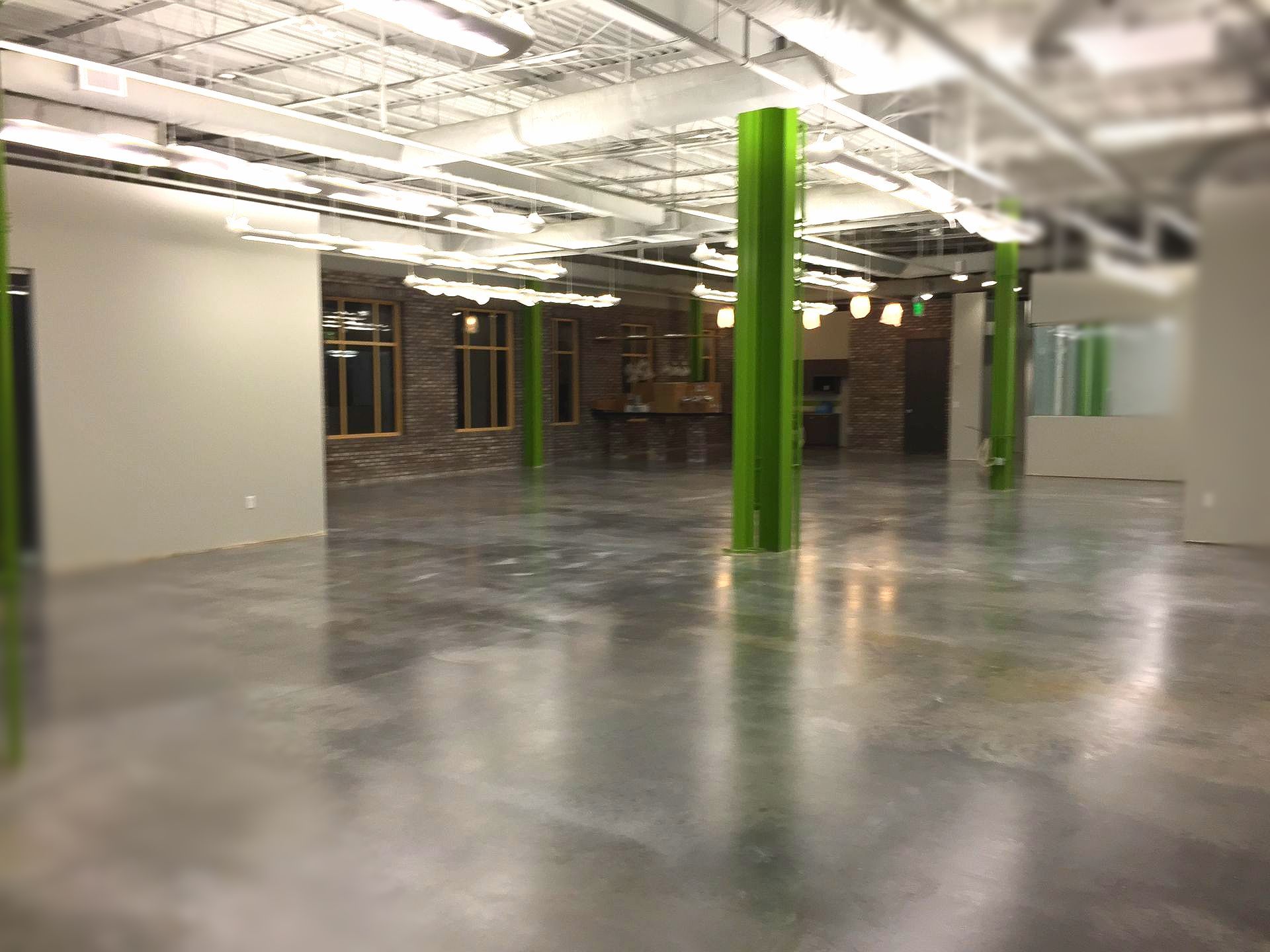 Spacious, empty room with polished concrete floor and green support columns. Exposed brick wall and overhead lighting present.