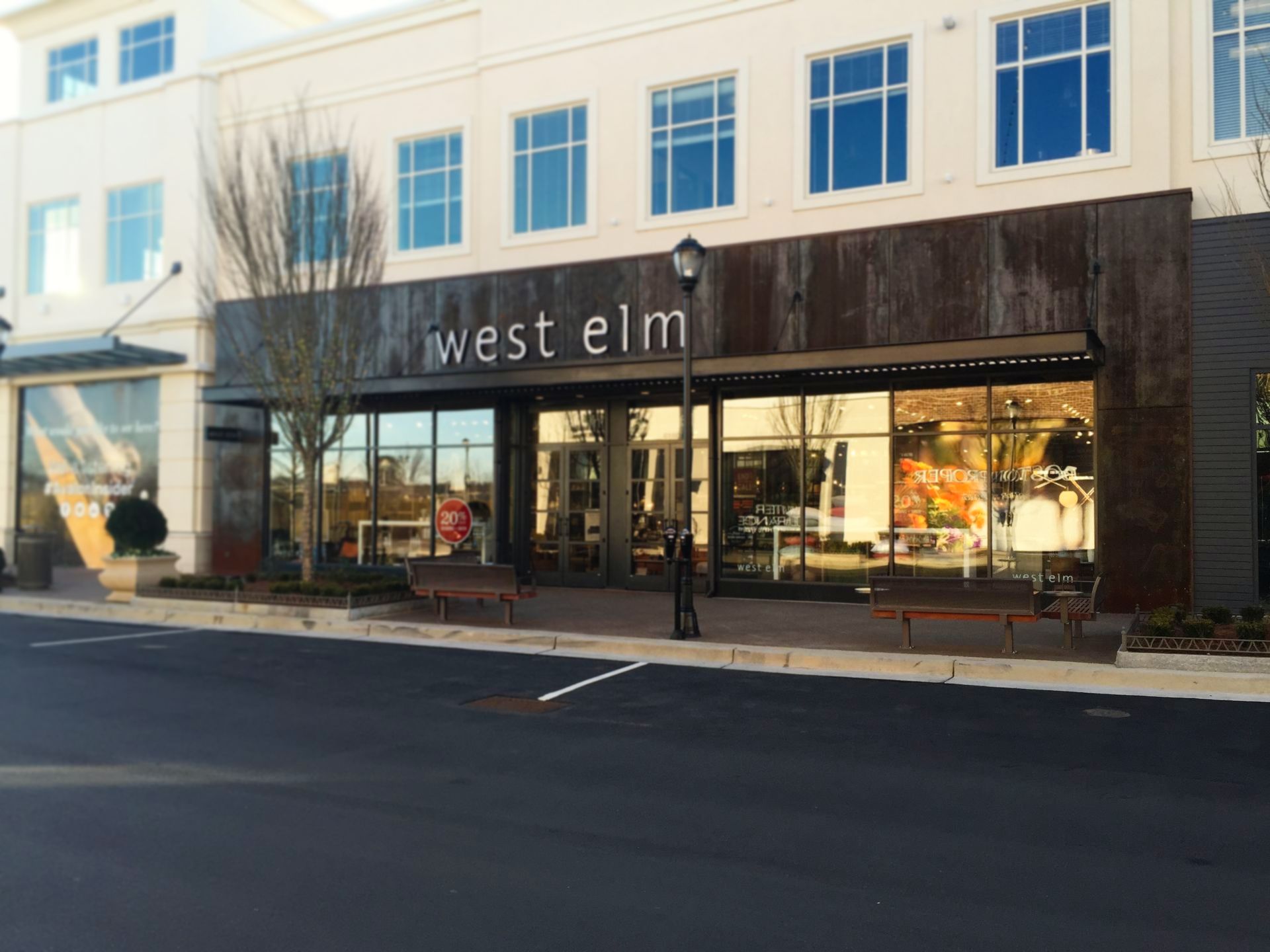 West Elm store exterior with large windows, a rusted metal awning, and the store name displayed. The building is situated in a shopping area with an asphalt parking lot.