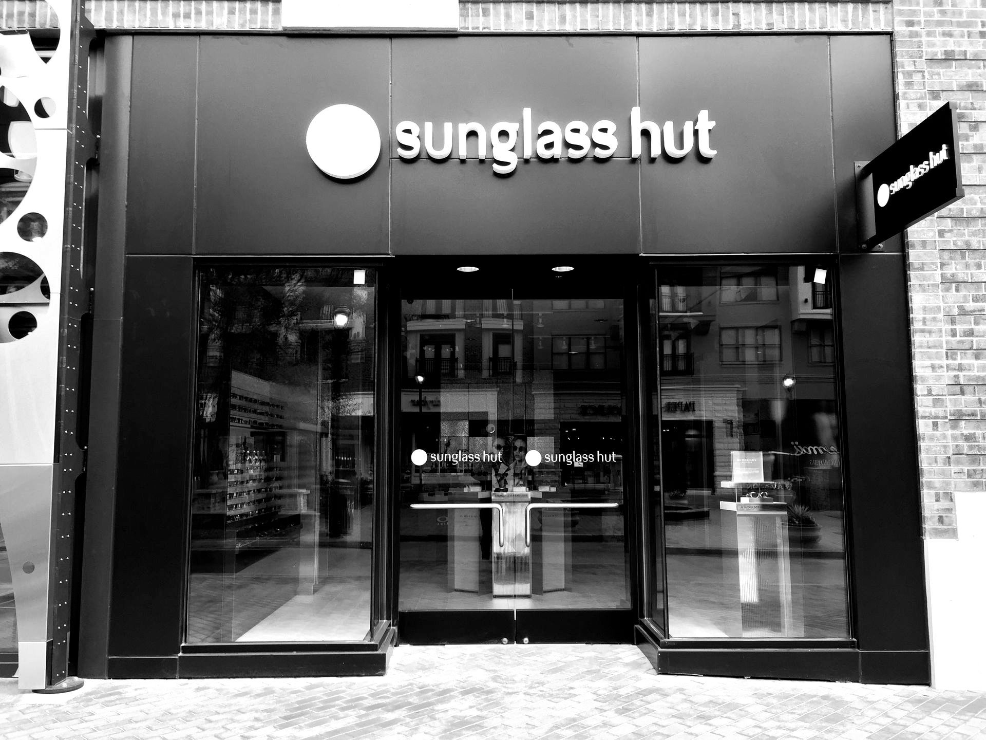 Black and white storefront of a Sunglass Hut. Glass front doors, sign above the door, and display cases visible inside.