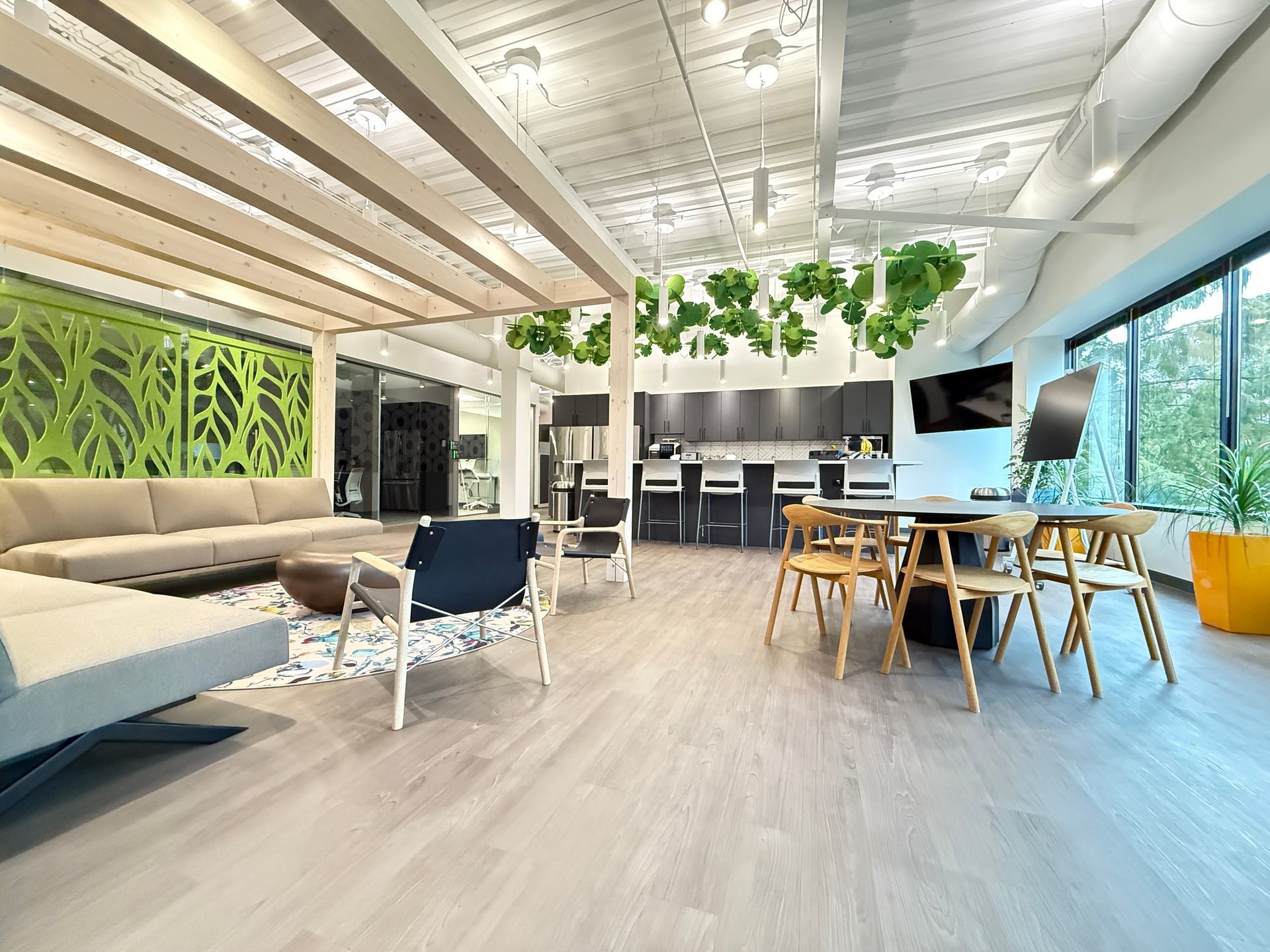 Spacious modern office interior with lounge area, kitchen, and dining table. Features include a green leaf-patterned wall, light wood flooring, and hanging plants.