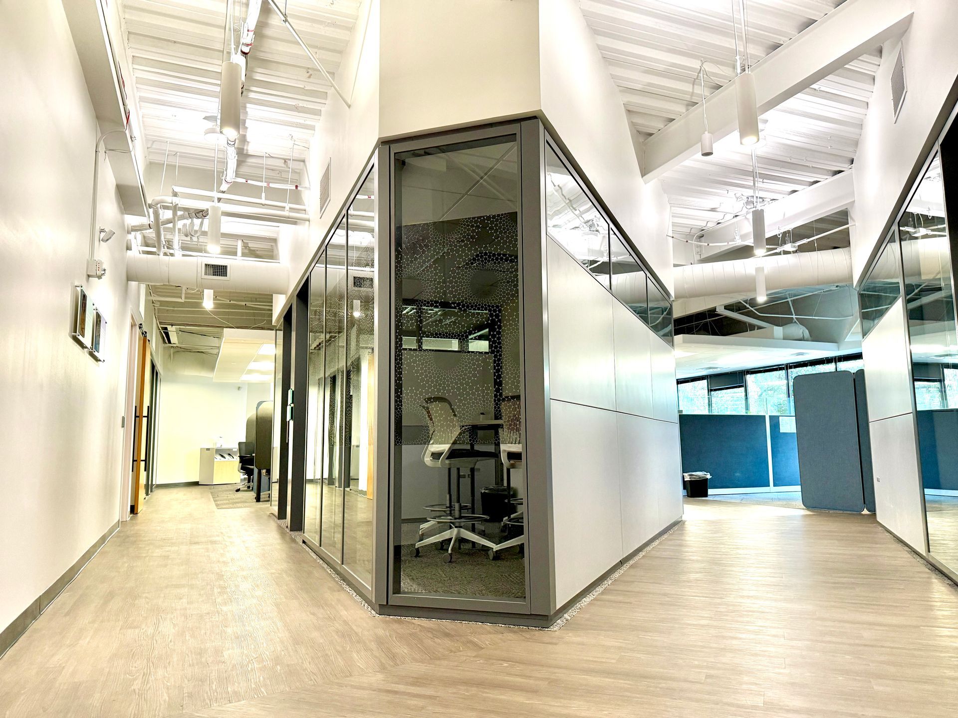 A modern office hallway with glass-walled rooms and white walls. Fluorescent lighting and exposed ductwork are visible on the ceiling.