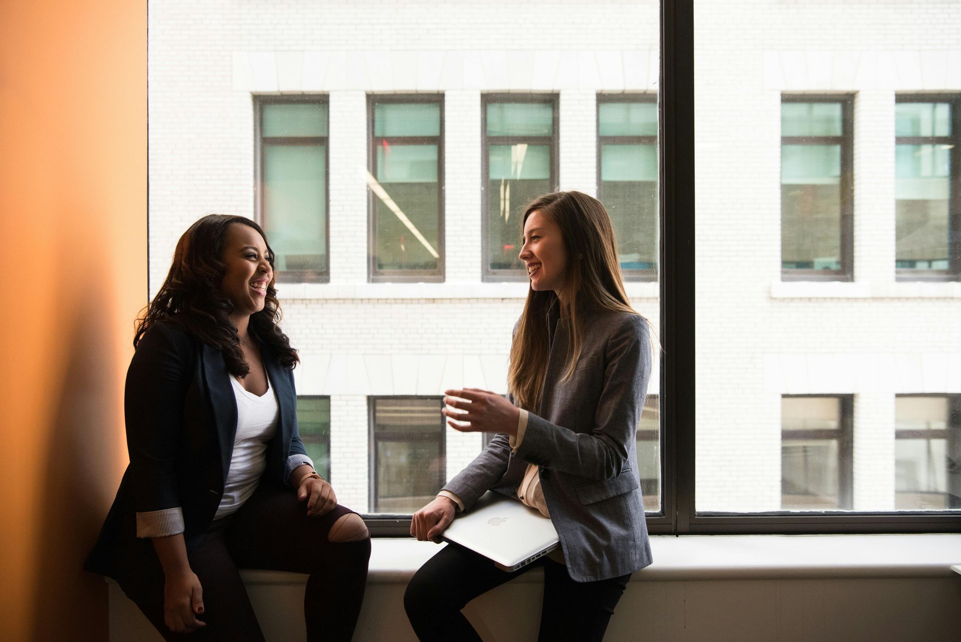 Two women laughing while sitting by a window. One holds a tablet, both in business attire. Orange wall background.