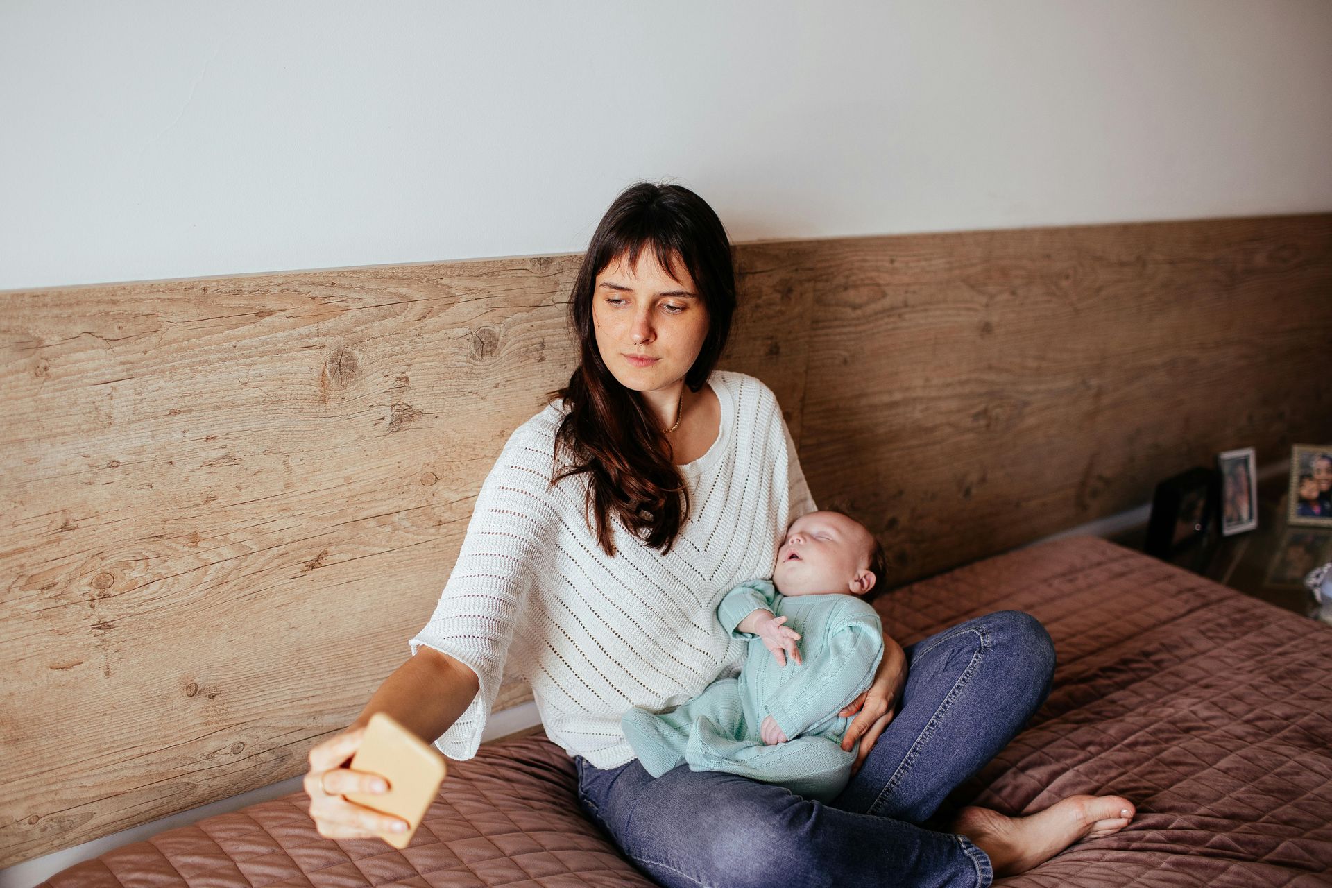 A person in a white sweater sitting on a bed, holding a baby in a teal outfit while taking a selfie.