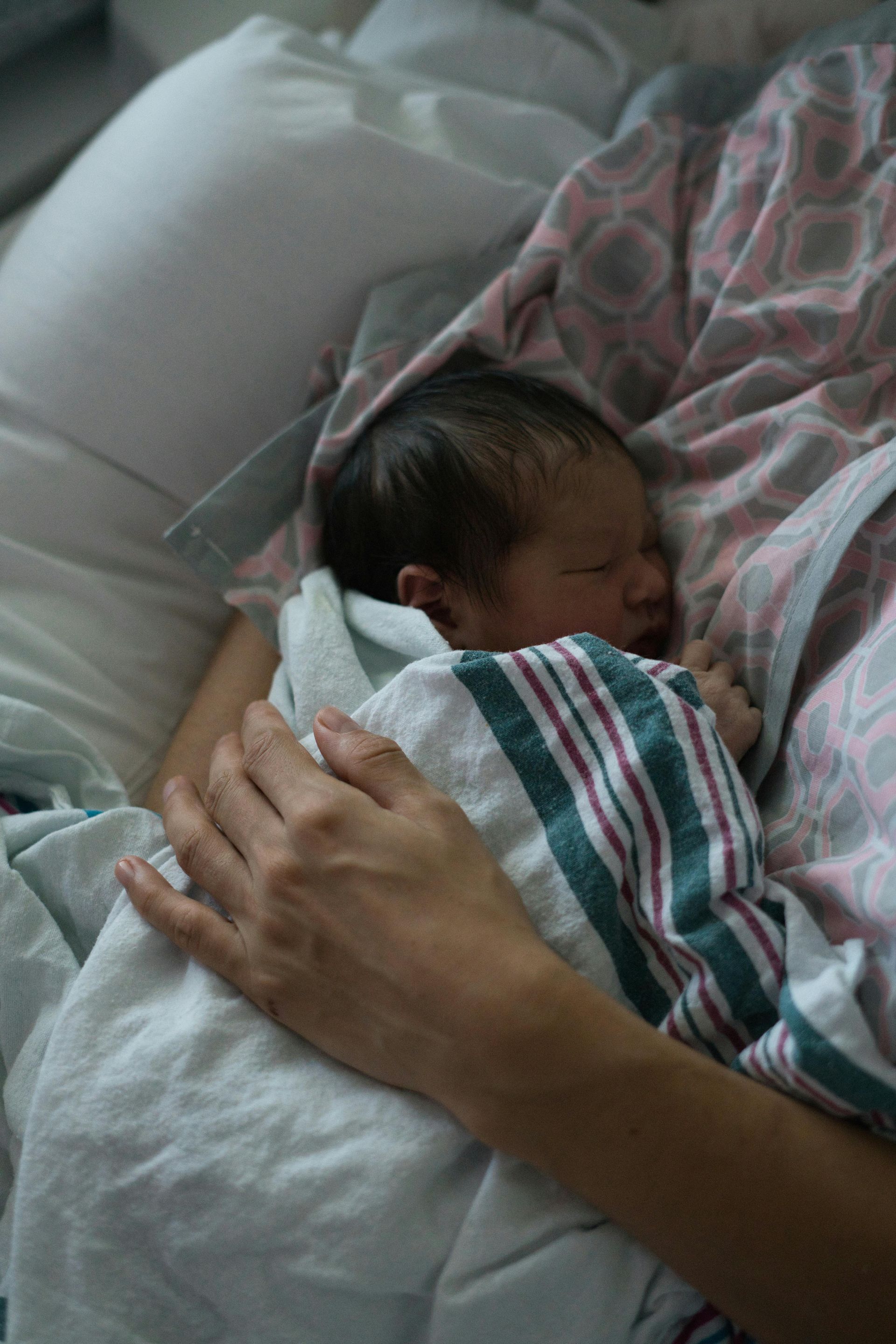 A newborn baby wrapped in a hospital blanket rests against a person's chest, held securely by their hand.