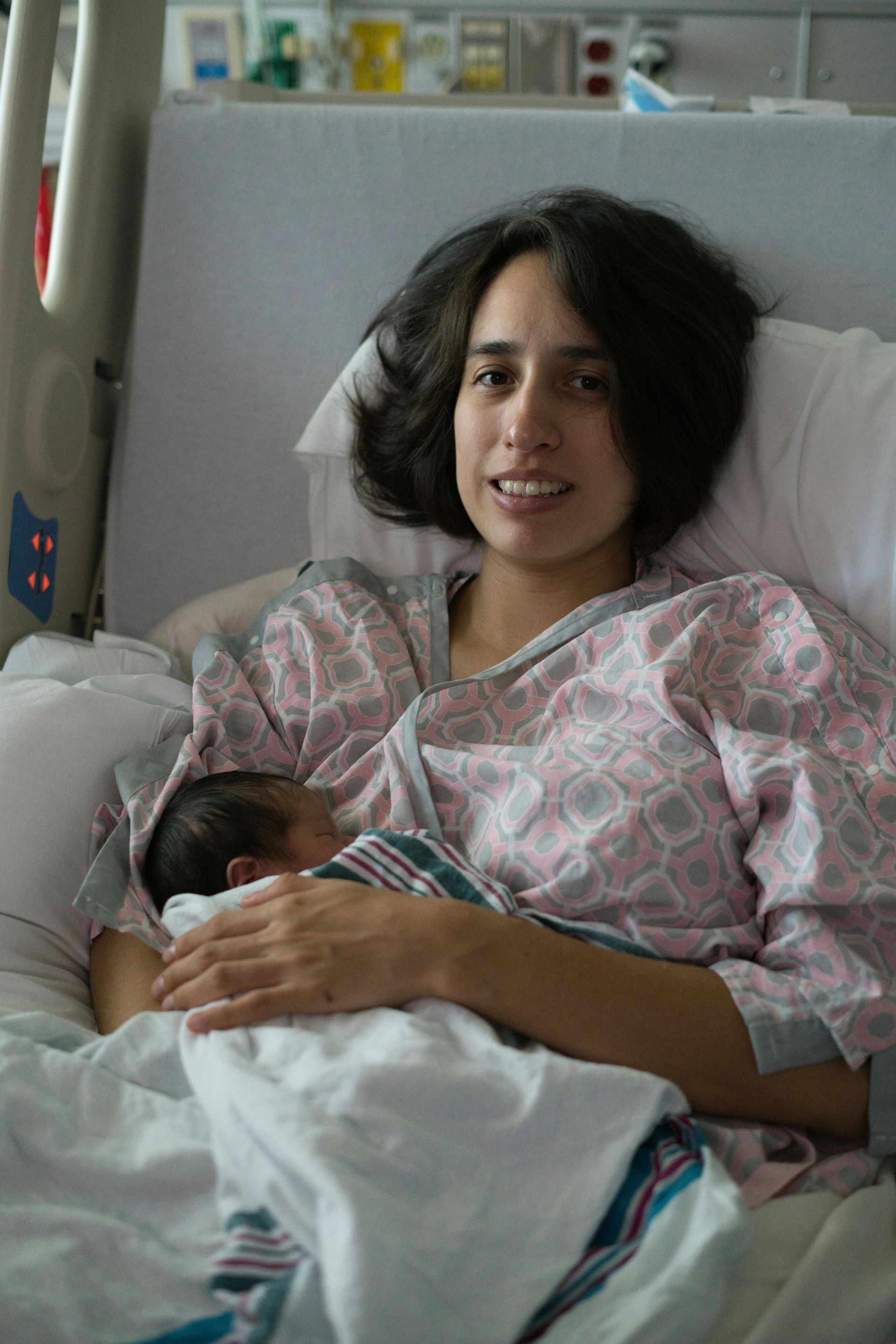 A person wearing a hospital gown rests in a hospital bed, looking at the camera while holding a newborn infant.