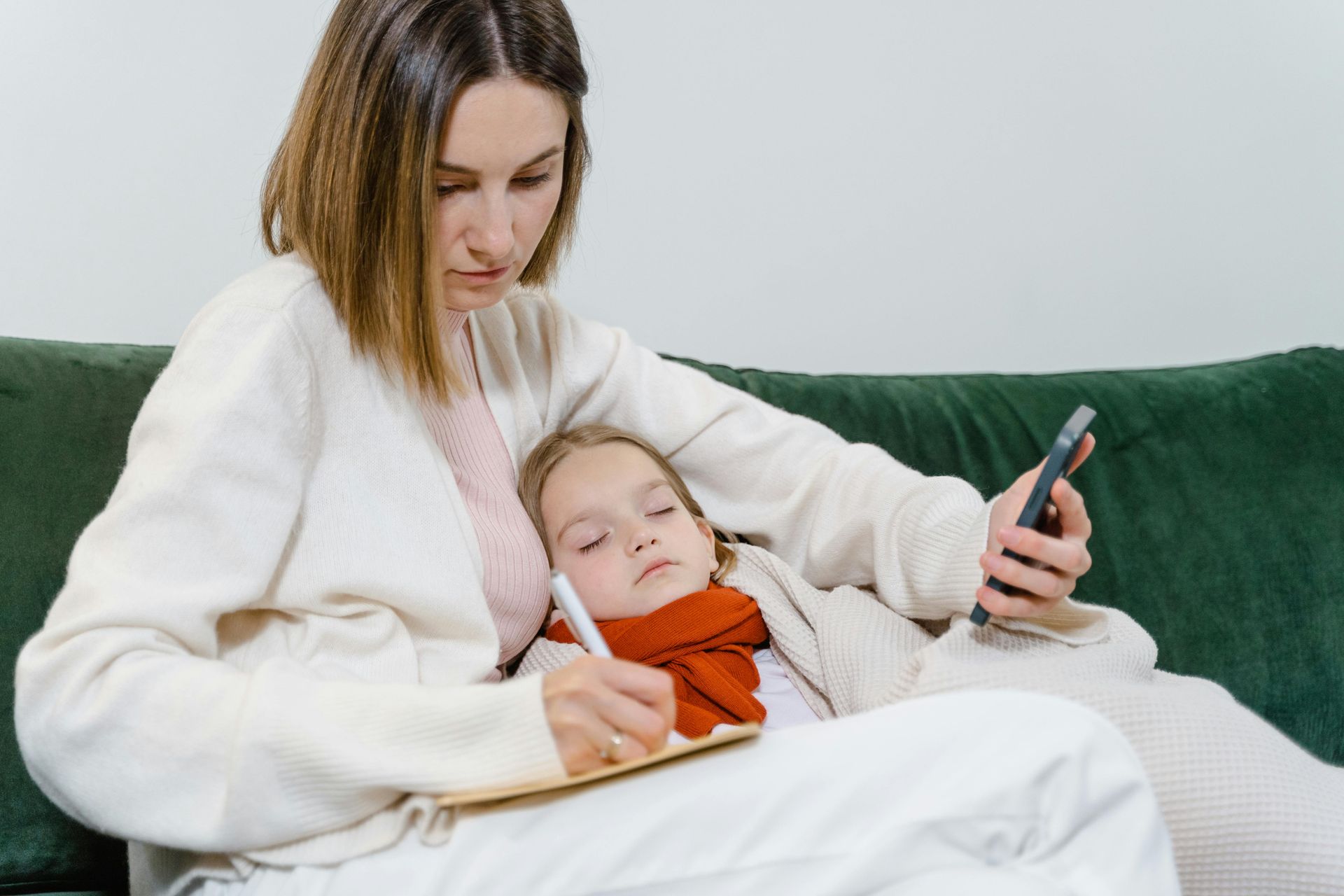 A person writes in a notebook on a green couch while holding a phone, with a child resting against them.