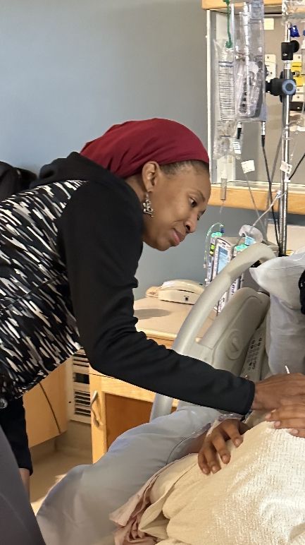 Woman in burgundy headscarf leans over, touching someone's arm in a hospital room.