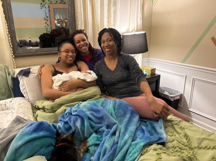 Three women smile in a bedroom. One holds a baby. Bed covered in blankets, dog visible.