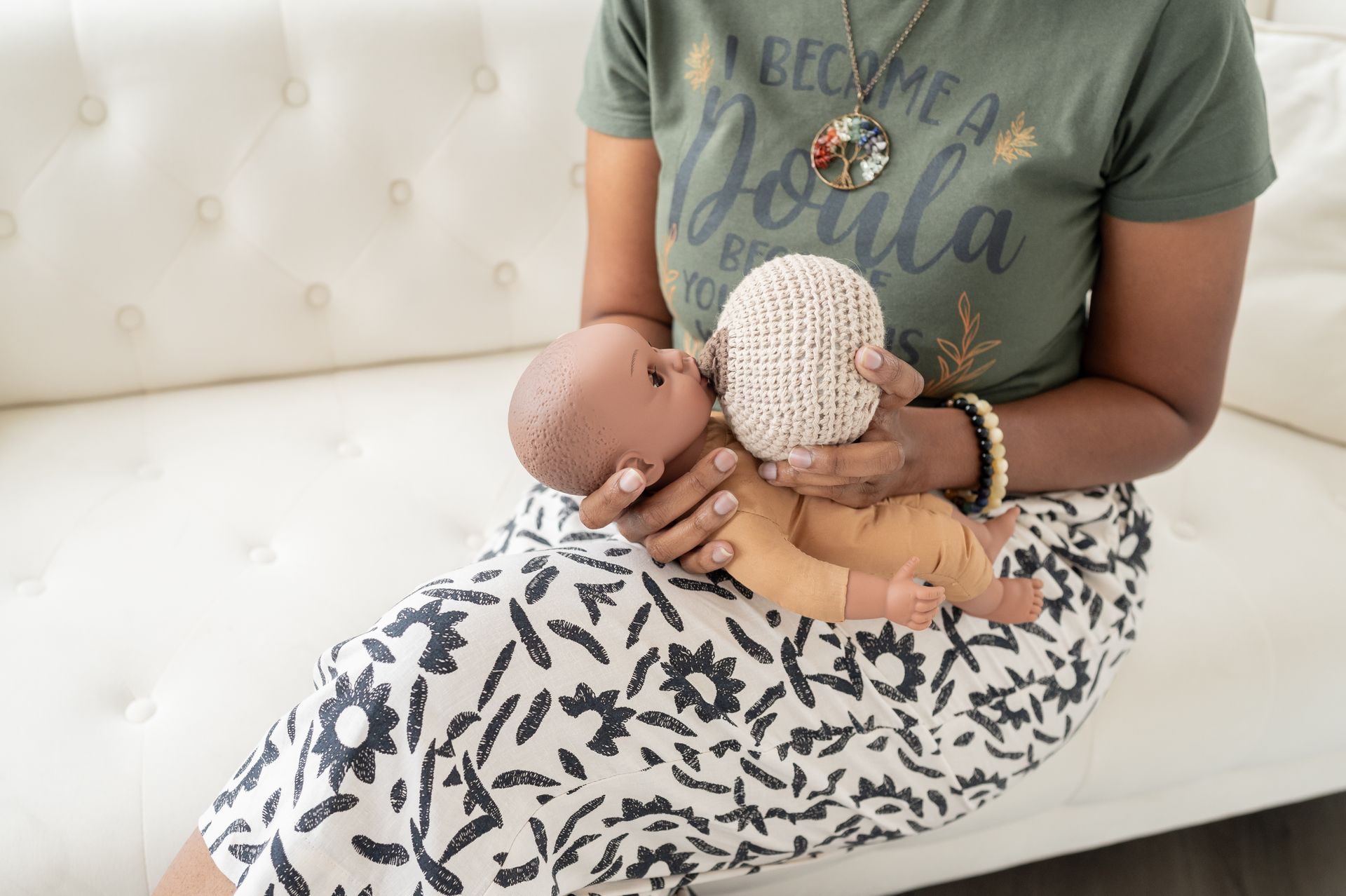 Woman in green shirt, patterned skirt holding a baby and a knitted breast model on a white couch.