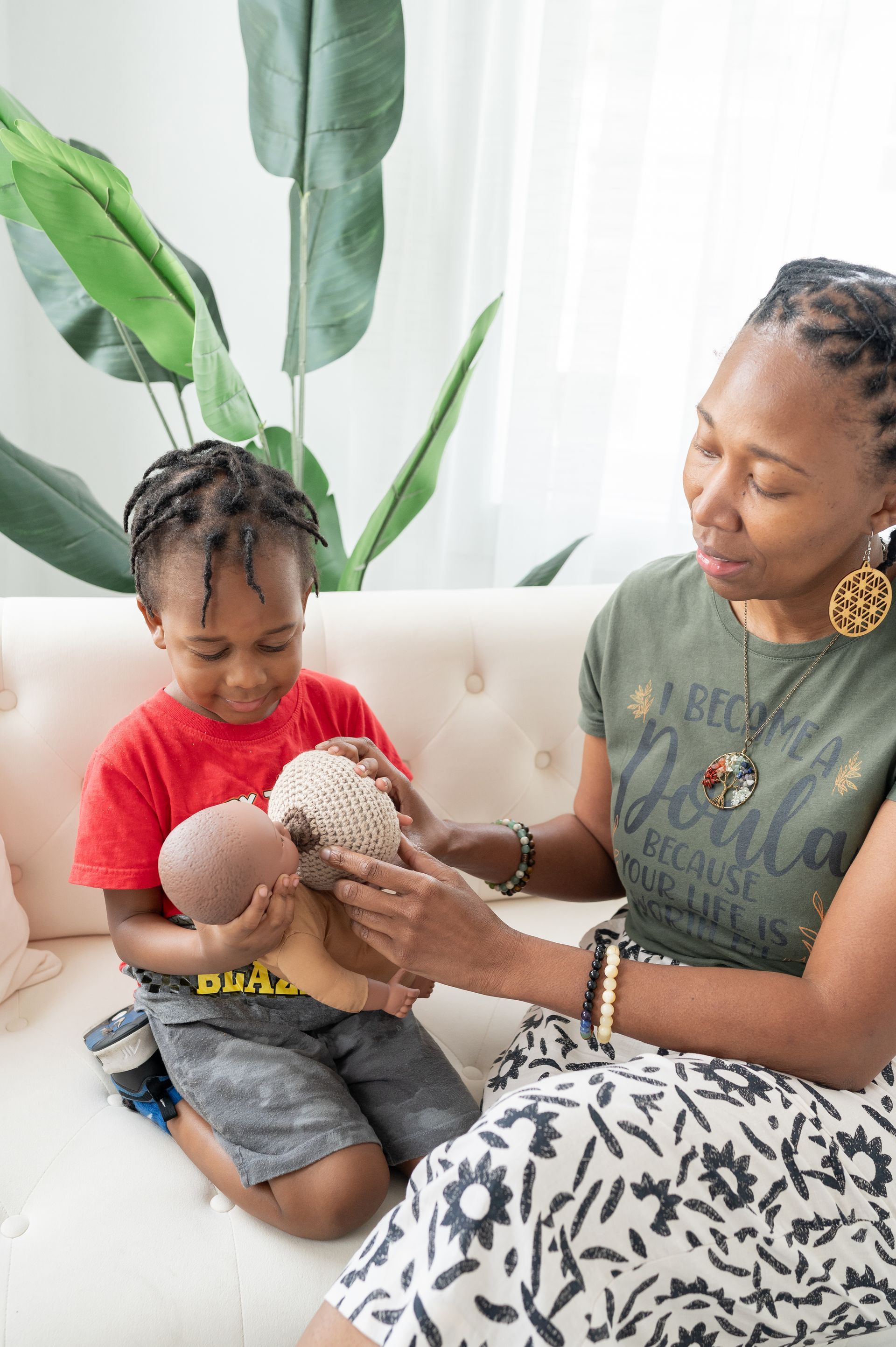Woman and child on couch, playing with doll. Child smiles, woman touches doll's head, indoor setting.