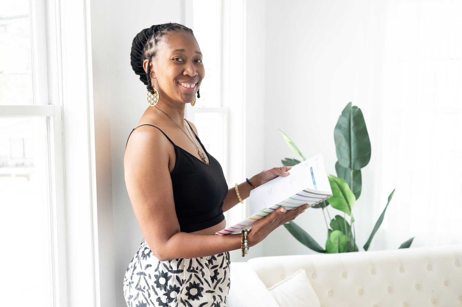 Woman smiling, holding binder near a window and plant, wearing a black top and patterned pants.