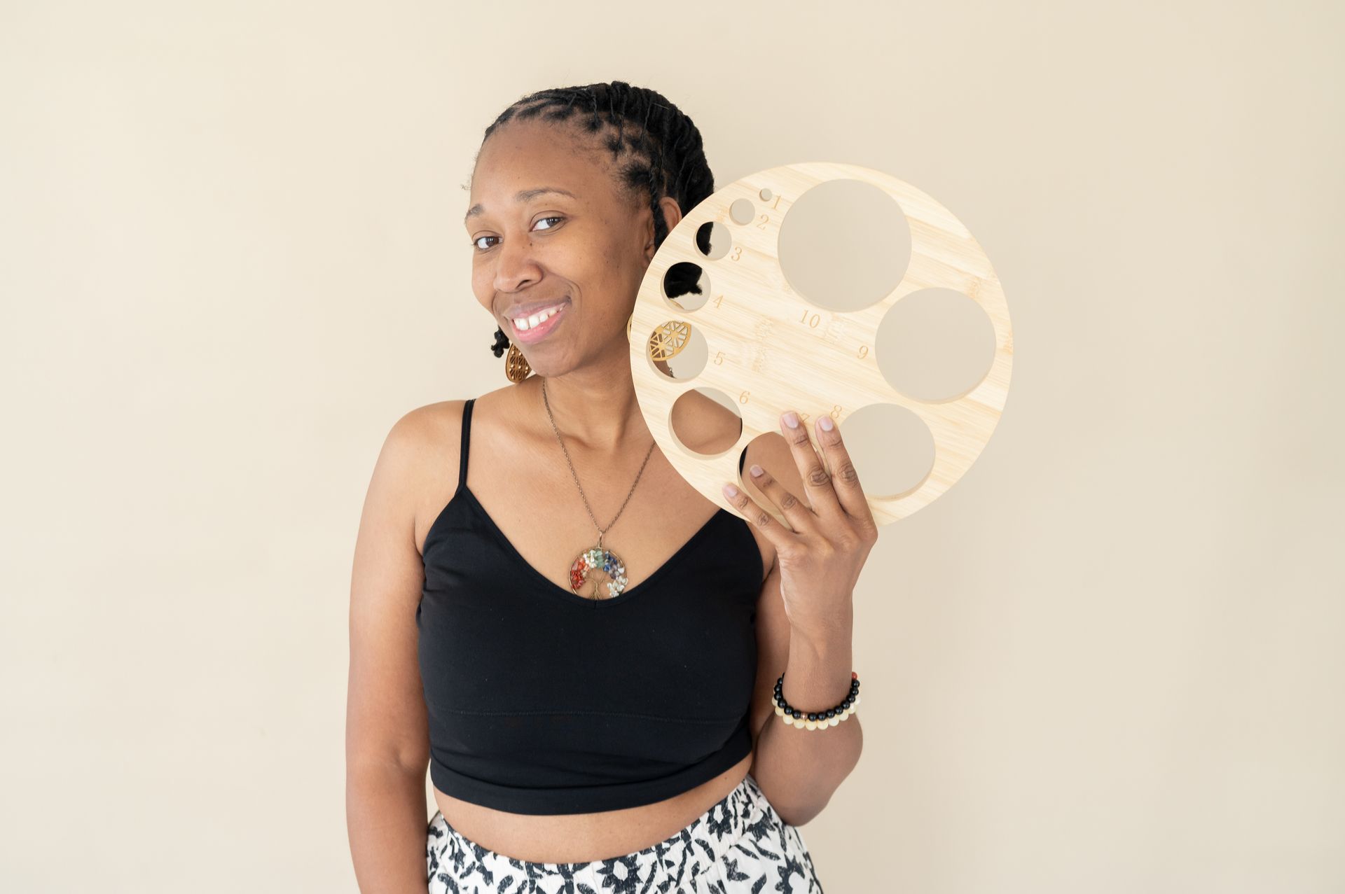 Woman holding a wooden, circular serving tray with cut-out sections; smiling in front of a tan wall.