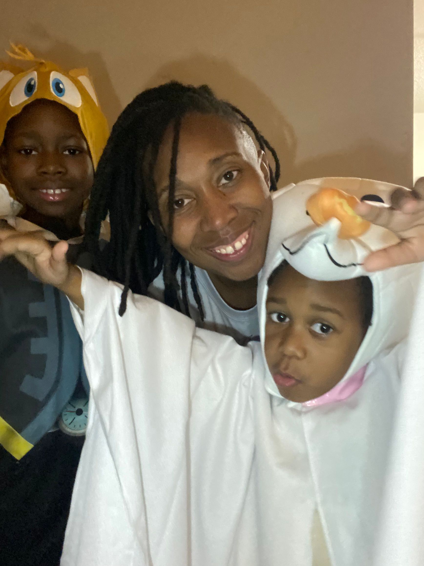 Woman smiles between two children in costumes, one with a ghost costume and the other in a character hat.