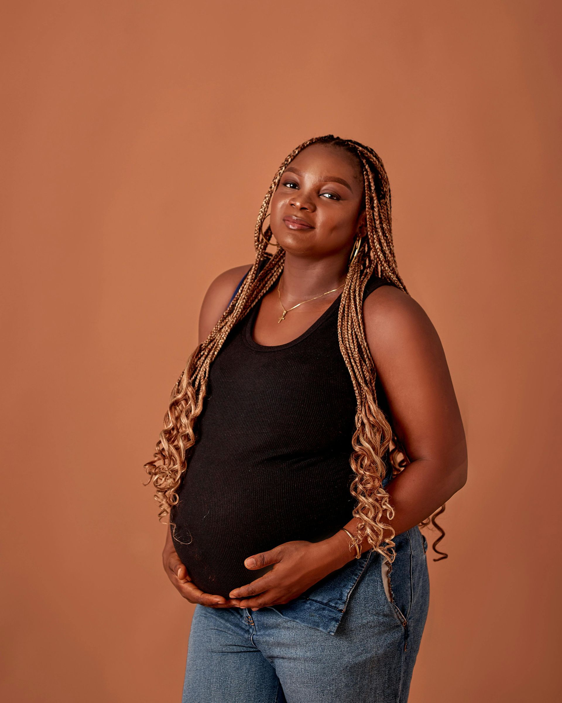 Pregnant person with long braided hair, wearing black top, jeans, hand on belly, brown background.