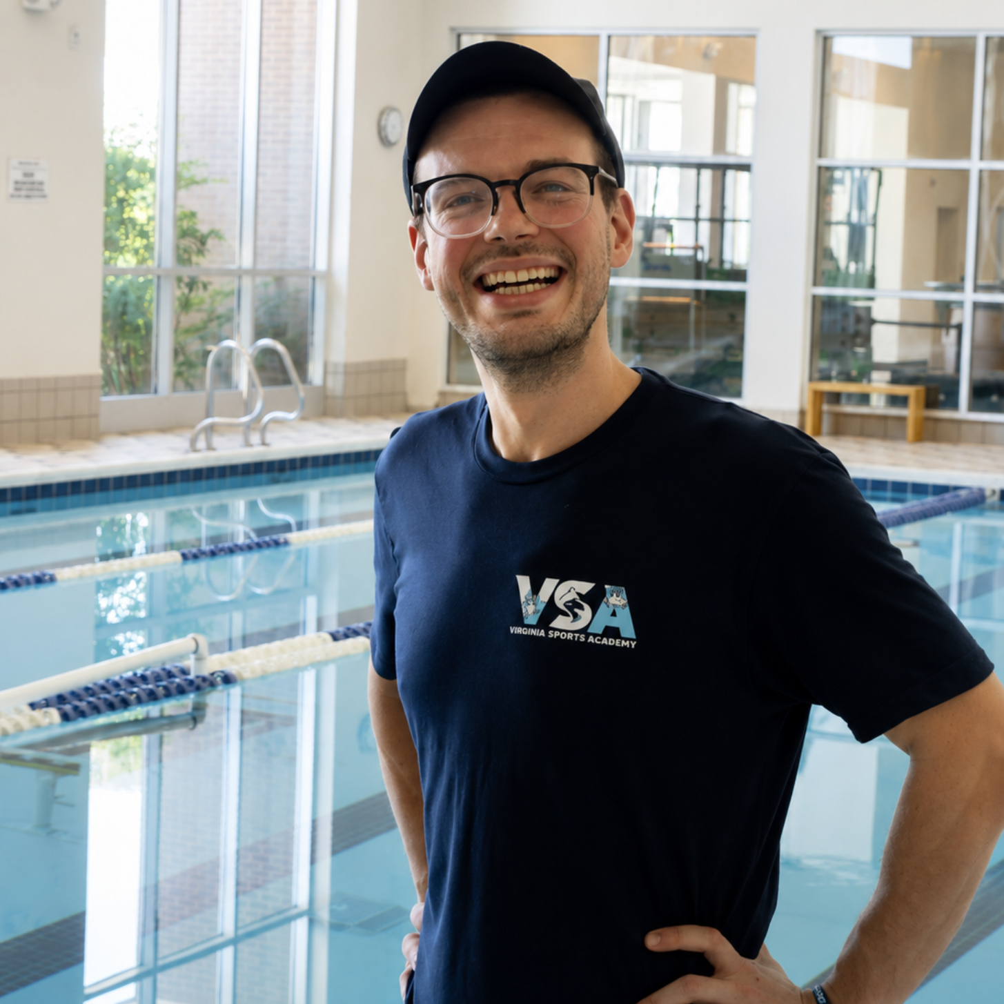 Smiling man in black cap and shirt standing by an indoor swimming pool