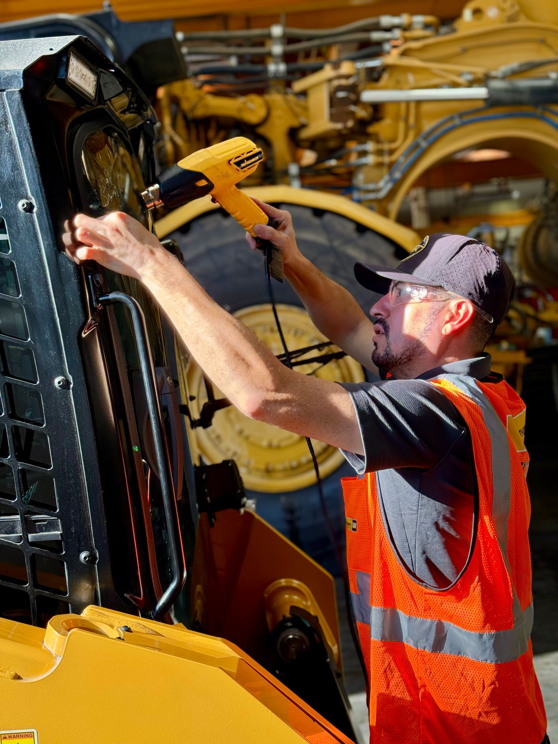 Mechanic using a heat gun on heavy machinery, wearing safety vest and cap. Yellow and black colors dominate.