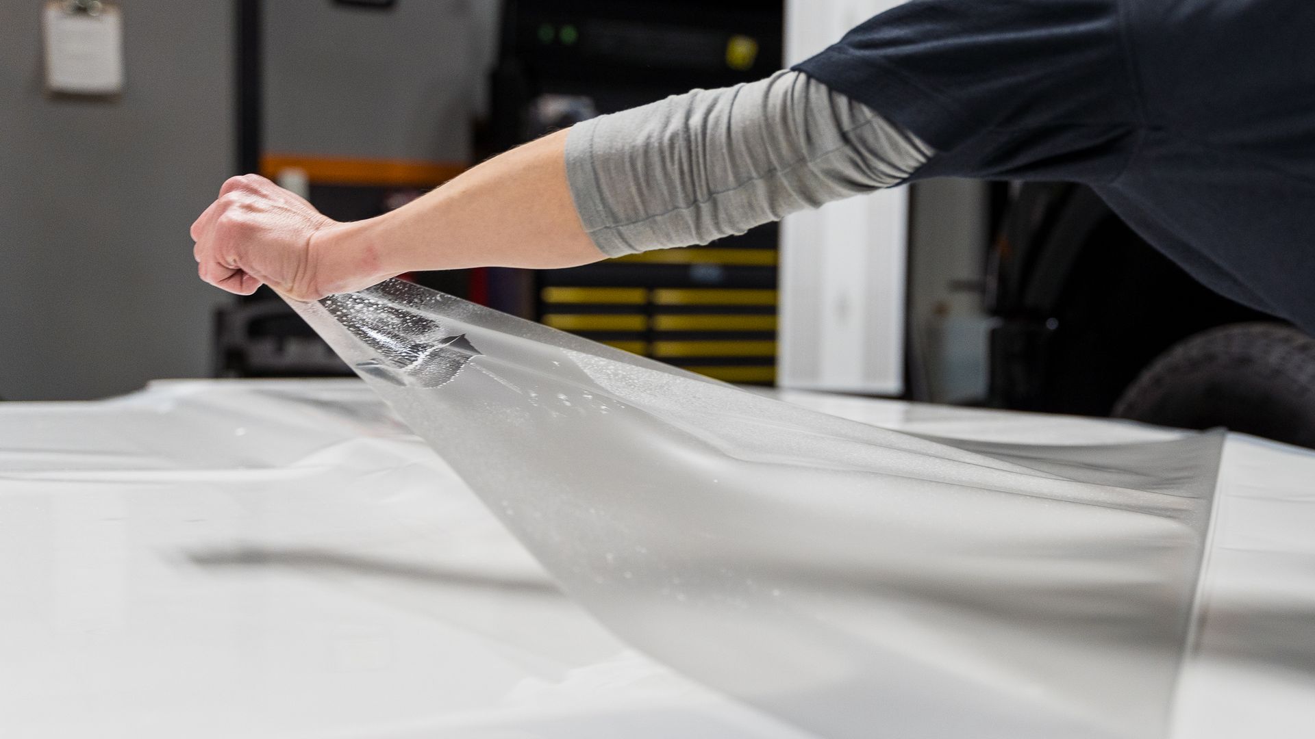 Person peels protective film from a white car hood in a shop.