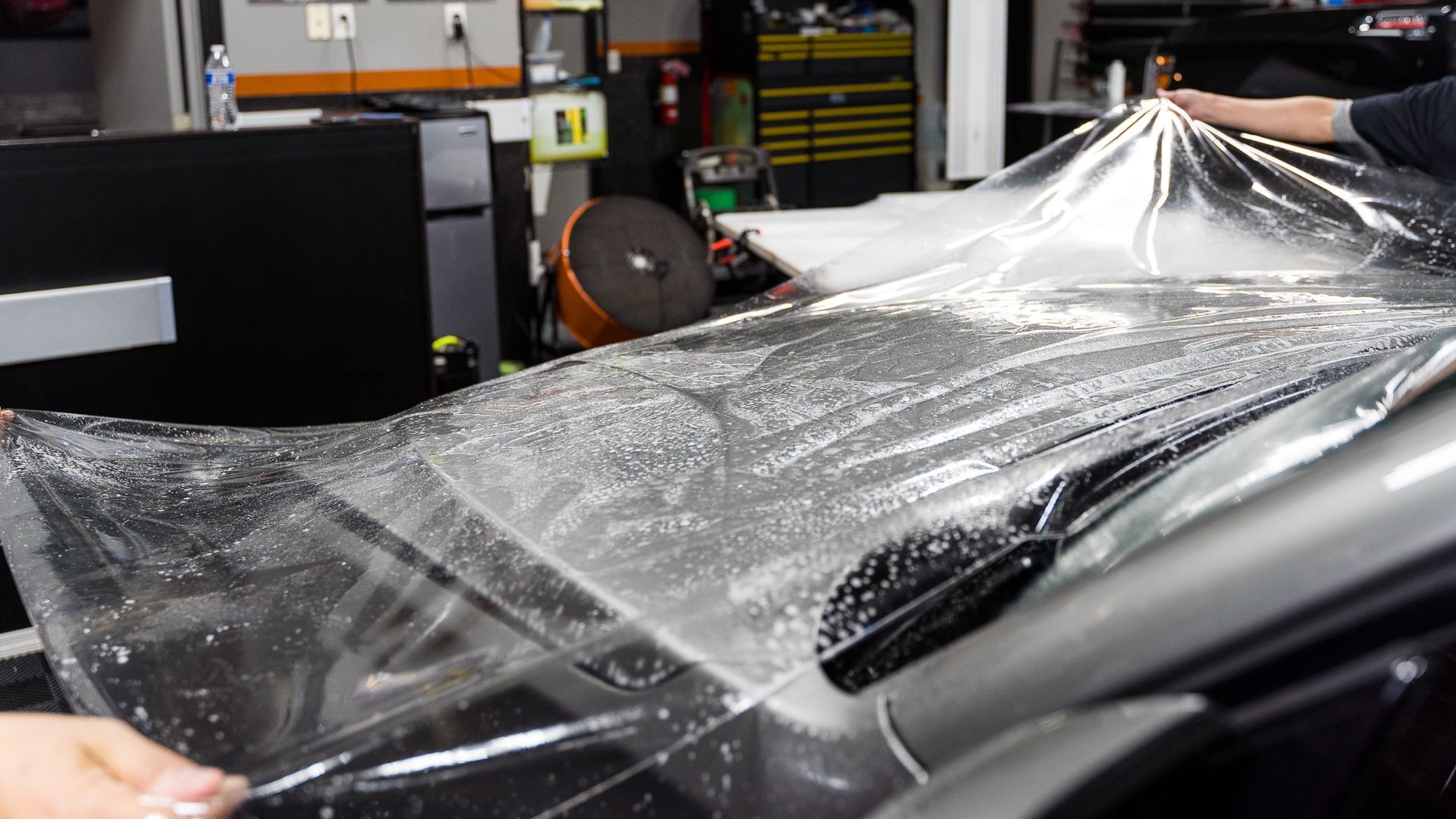 Two people applying clear protective film to a car hood in a garage.