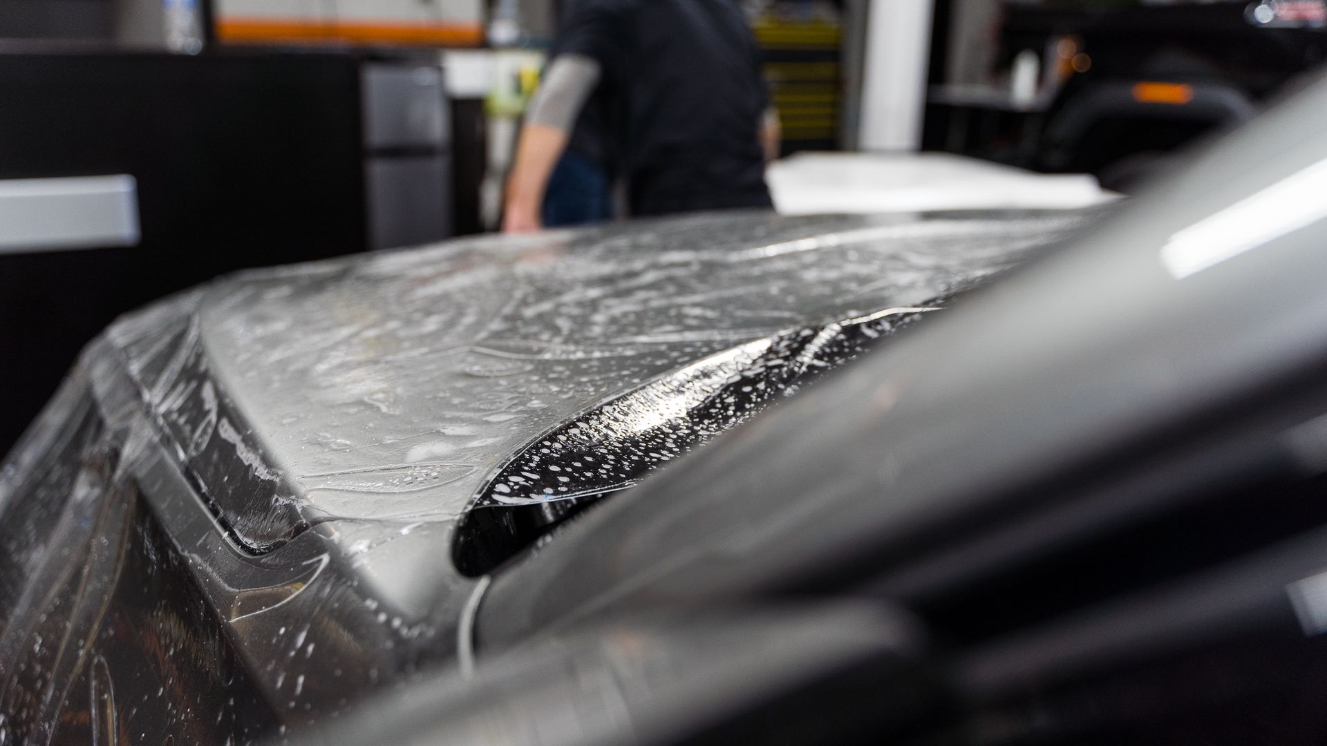 Car hood covered in protective film, in a shop setting, with a person in the background.