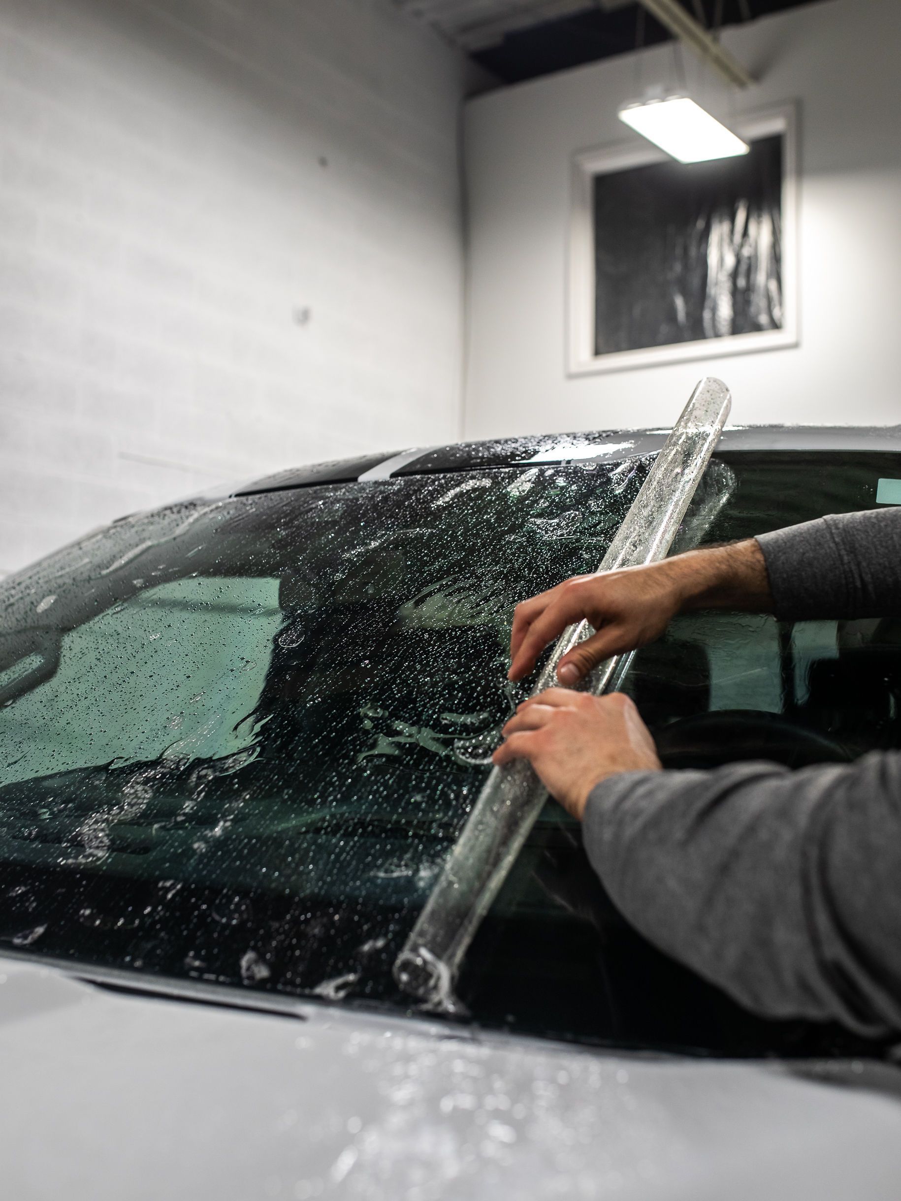 A person uses a squeegee to smooth window tint onto the wet windshield of a car in a workshop.