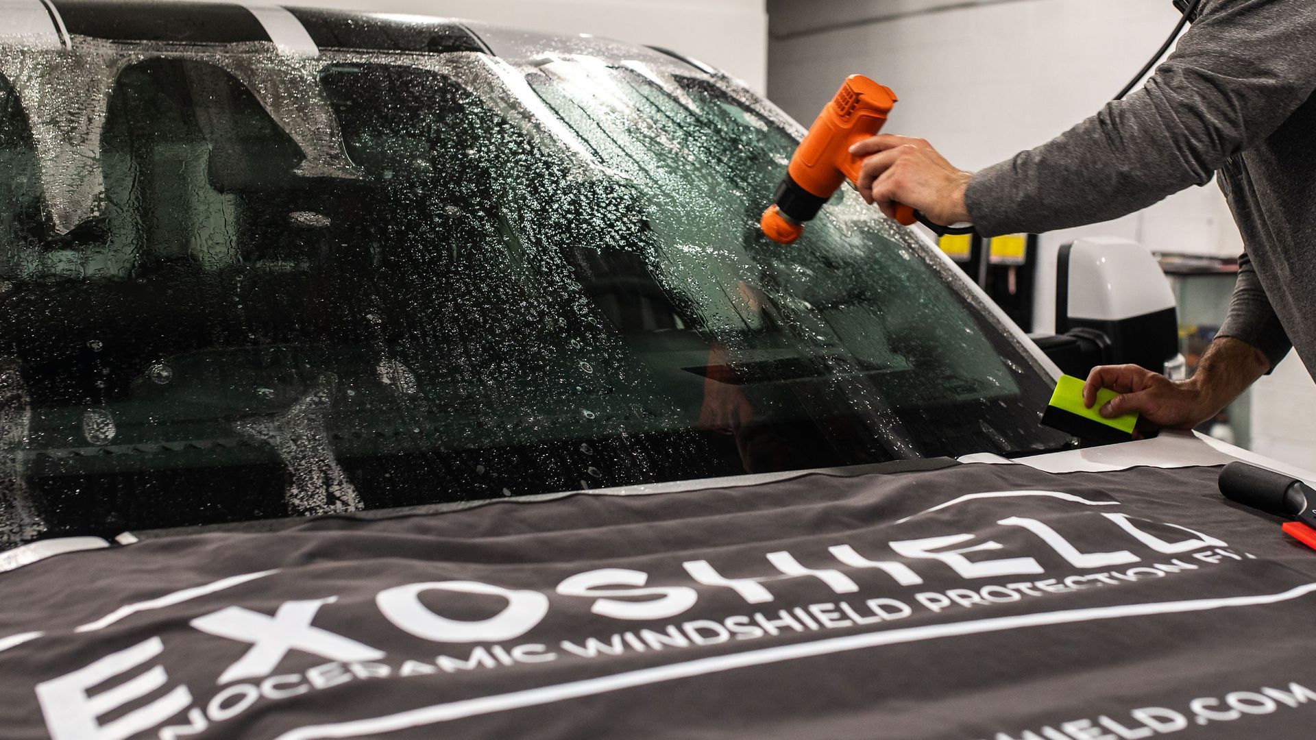 A technician uses a heat gun to apply protective film to a vehicle windshield over a branded Exoshield mat.