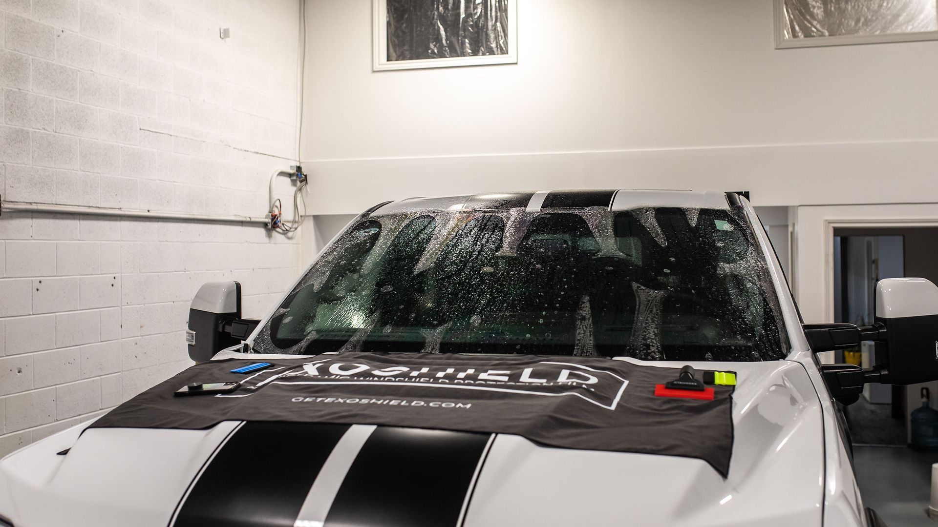 A white truck with black racing stripes in a garage, undergoing a windshield application with a protective black mat.