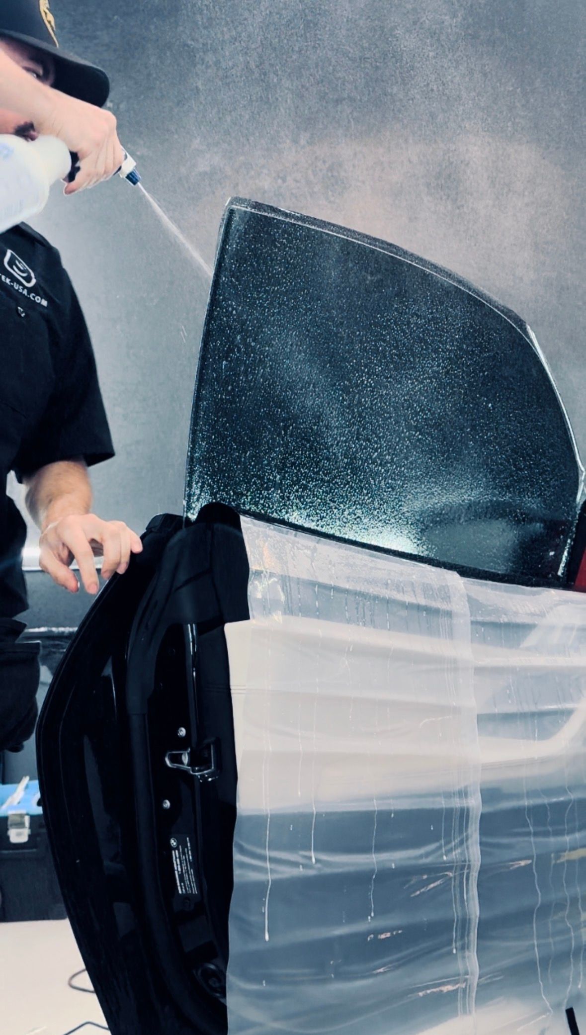 Person spraying a car window with a spray bottle, likely applying window film. Window is partially covered in plastic.