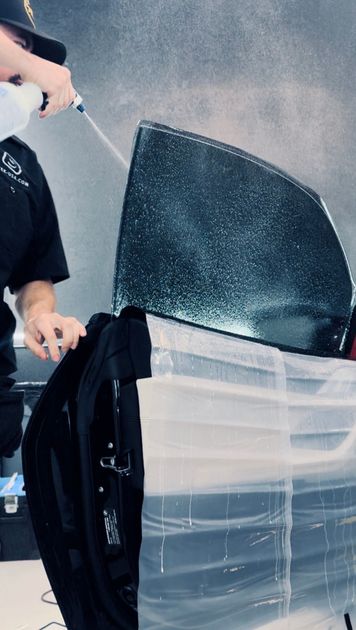 Person spraying a car window with a spray bottle, likely applying window film. Window is partially covered in plastic.