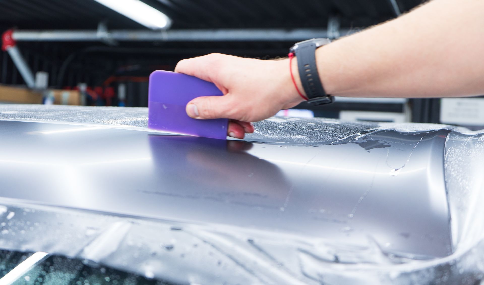 Two people applying clear protective film to a car hood in a garage.