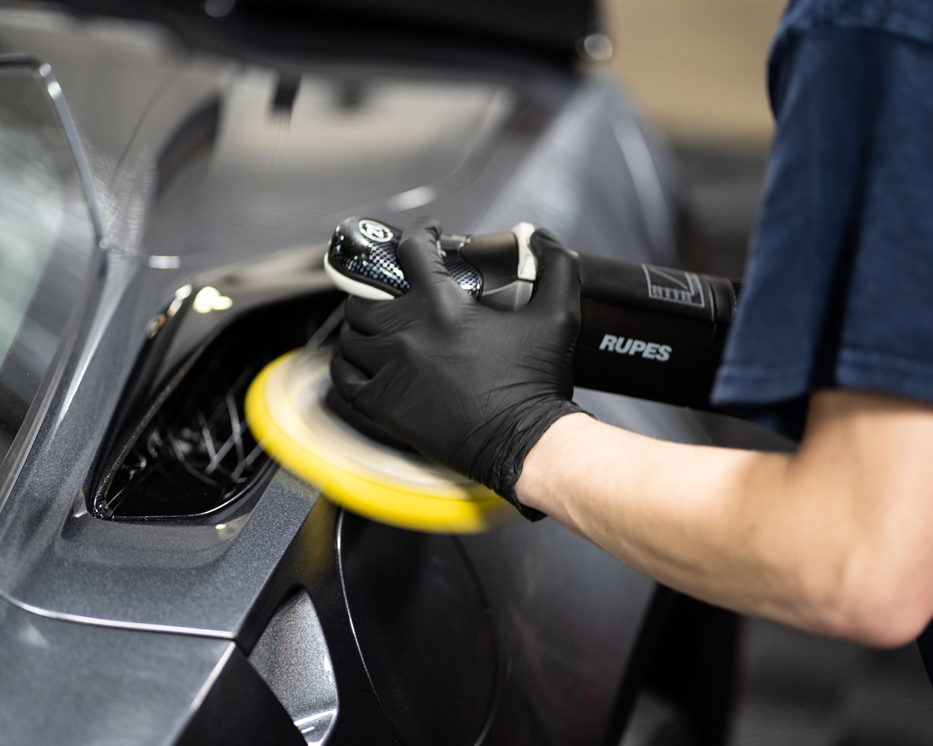 Person wearing black gloves polishing a gray car with a Rupes polisher, focusing on a vent.