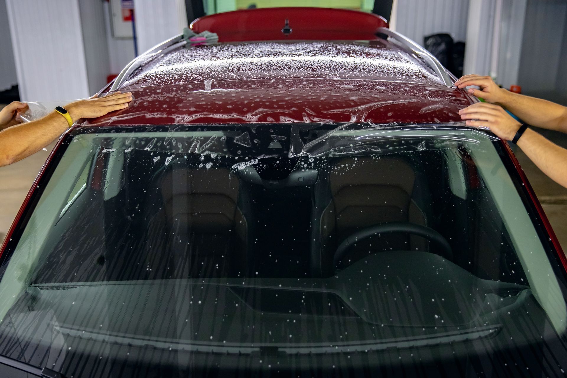 People applying a protective film to the roof and windshield of a red car in a garage.