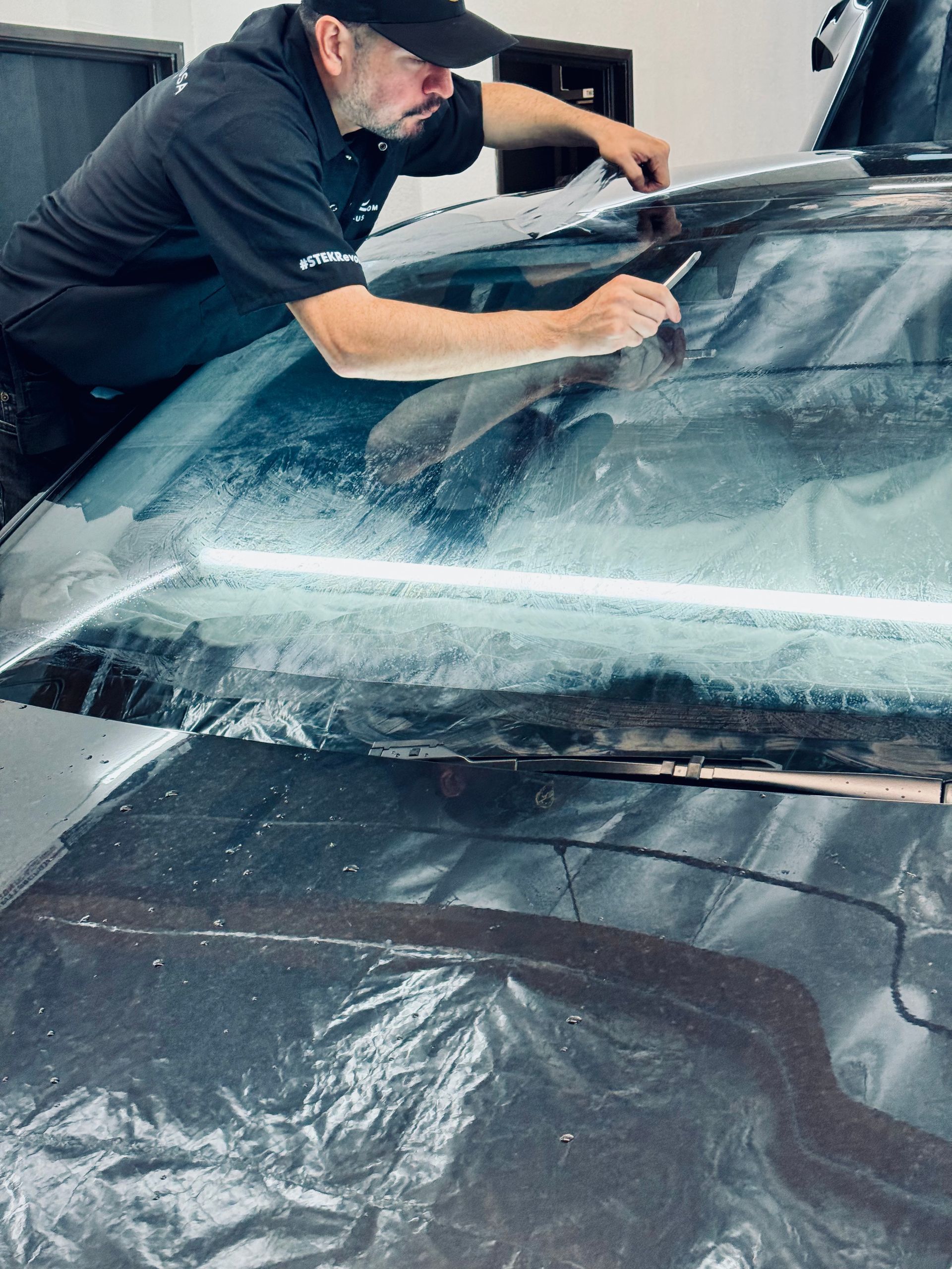 Man applying tint to a car windshield in a shop; he is kneeling, focused on his work.
