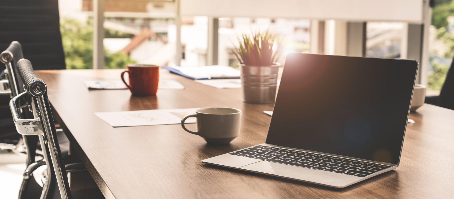 Laptop and two mugs on a wooden conference table in a bright room with a view of greenery.