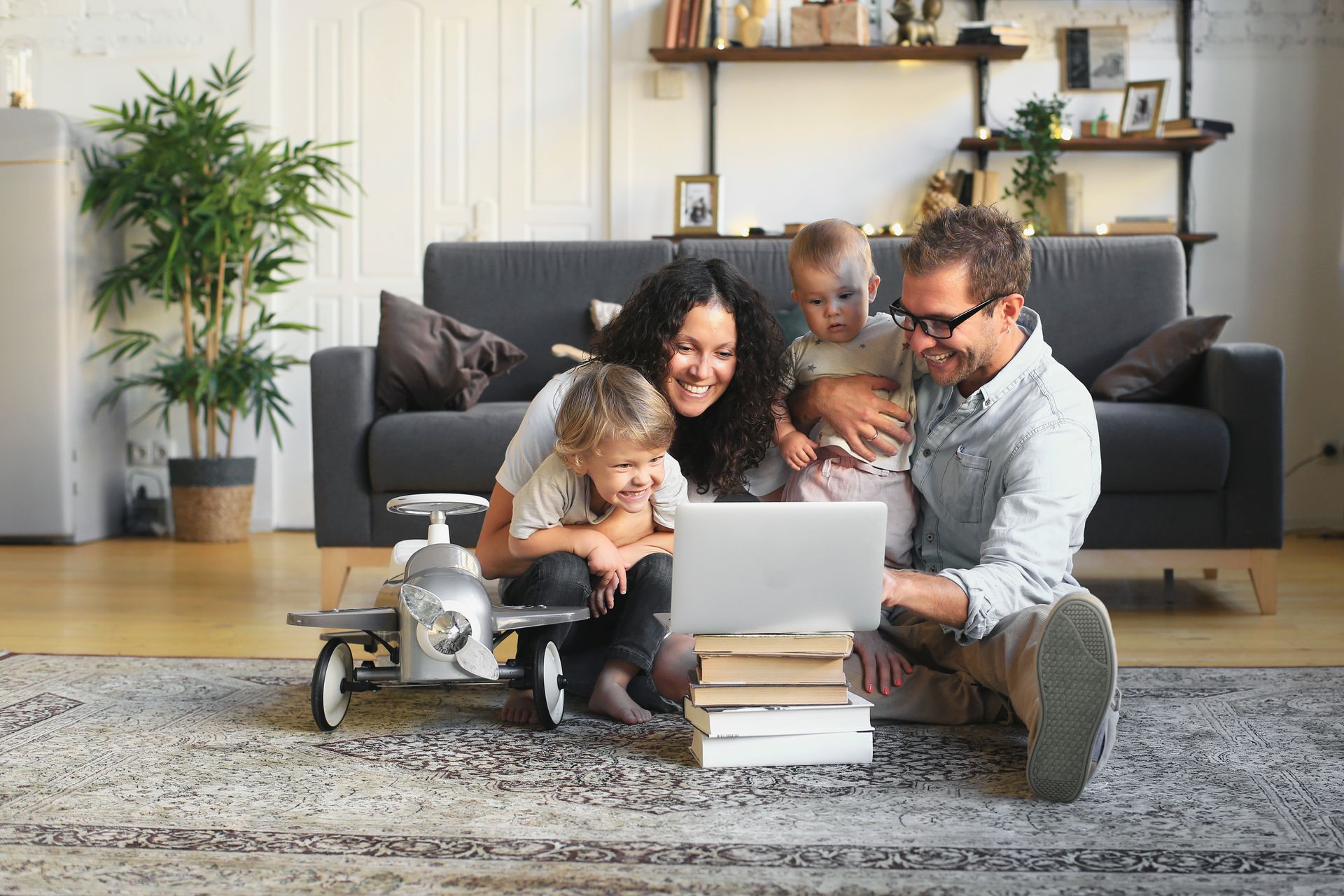 Family of four smiles while looking at a laptop. They are in a living room with a rug and couch.