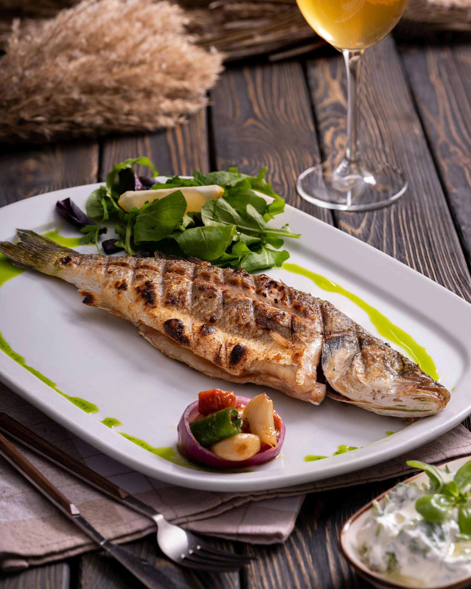 Gegrillter Fisch auf einem weißen Teller, mit Salat, eingelegtem Gemüse und Weißwein auf einem dunklen Holztisch.