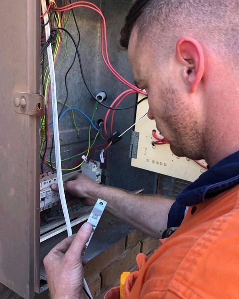 A Man in An Orange Shirt Is Working on An Electrical Box — Paton's Electrical Services in Hervey Bay, QLD