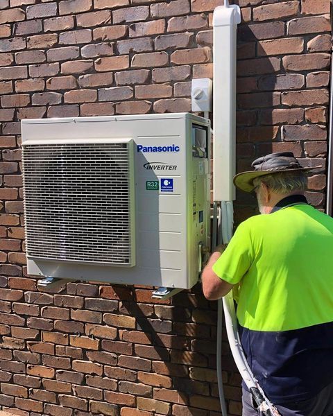 A Man Is Installing a Panasonic Air Conditioner on A Brick Wall — Paton's Electrical Services in Kawungan, QLD