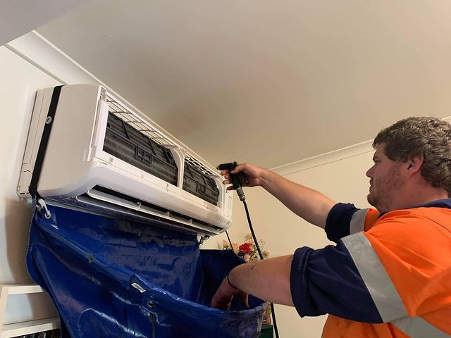 A Man Is Cleaning an Air Conditioner with A Hose — Paton's Electrical Services in K'Gari, QLD