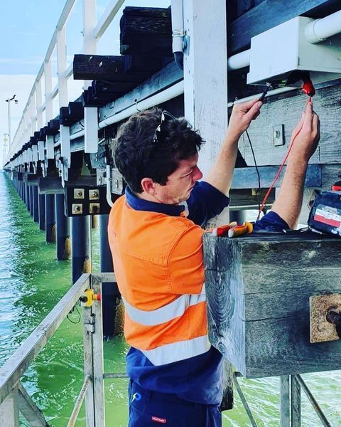 A Man in An Orange Shirt Is Working on A Bridge — Paton's Electrical Services in Hervey Bay, QLD
