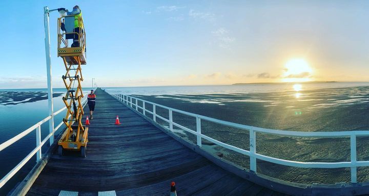 A Man Is Standing on A Scissor Lift on A Pier Overlooking the Ocean — Paton's Electrical Services in Urangan, QLD