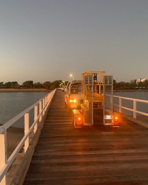 A Yellow Truck Is Driving Down a Wooden Bridge Over a Body of Water — Paton's Electrical Services in Pialba, QLD