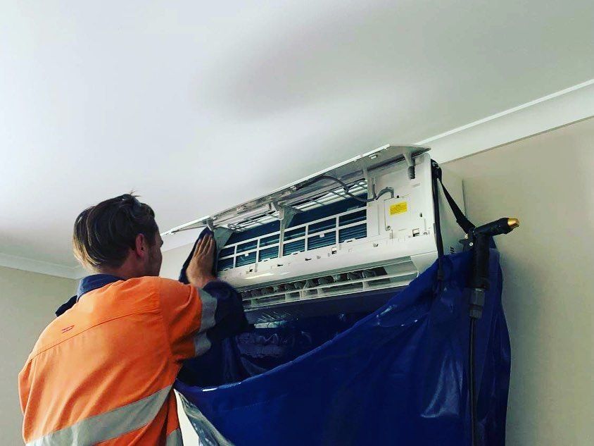 A Man Is Cleaning an Air Conditioner with A Blue Tarp — Paton's Electrical Services in Maryborough, QLD