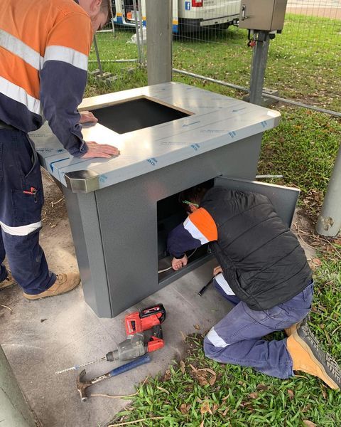Two Men Are Working on A Metal Box with A Hammer and Drill — Paton's Electrical Services in Maryborough, QLD