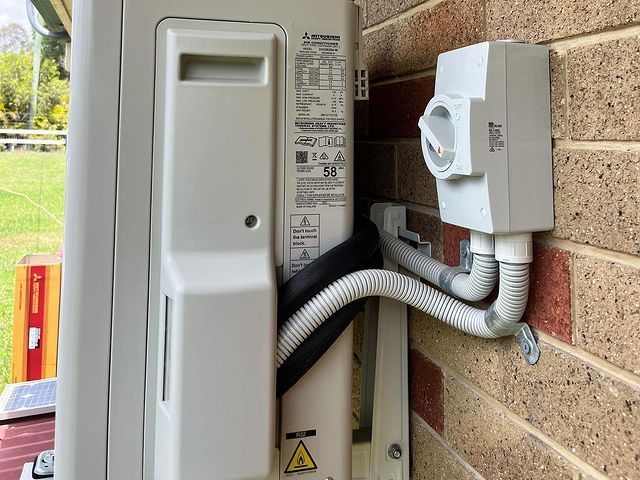A White Air Conditioner Is Attached to A Brick Wall — Paton's Electrical Services in K'Gari, QLD