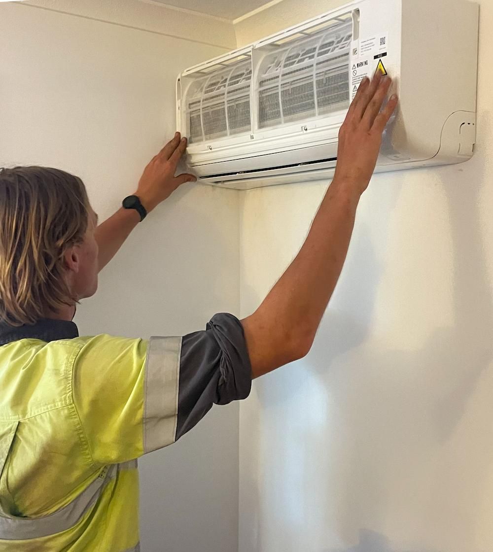 A Man is Cleaning an Air Conditioner on a Wall — Paton's Electrical Services in Urangan, QLD