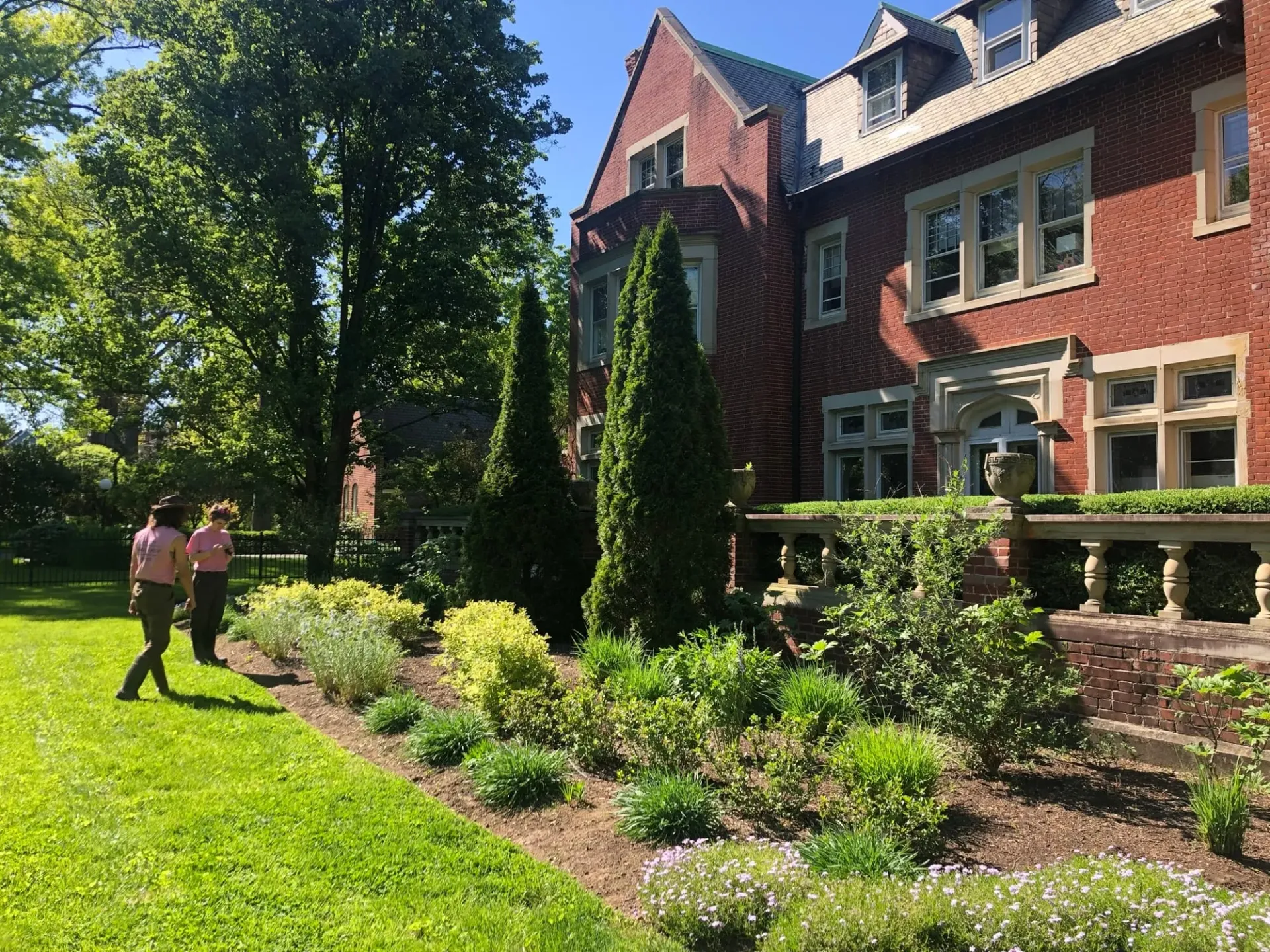 Two people in pink shirts look at a garden of various plants next to a red brick building.