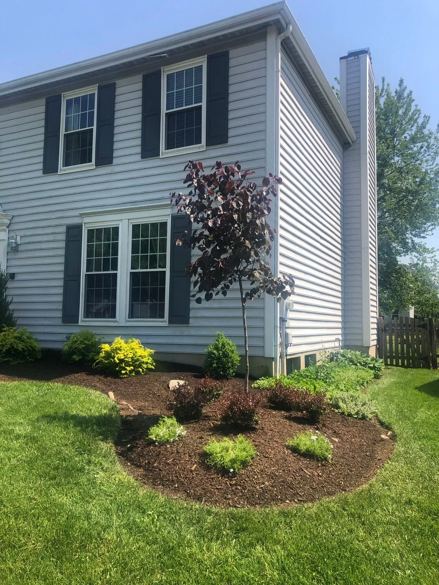 Two-story house with gray siding, black shutters, and a landscaped front yard with a small tree.