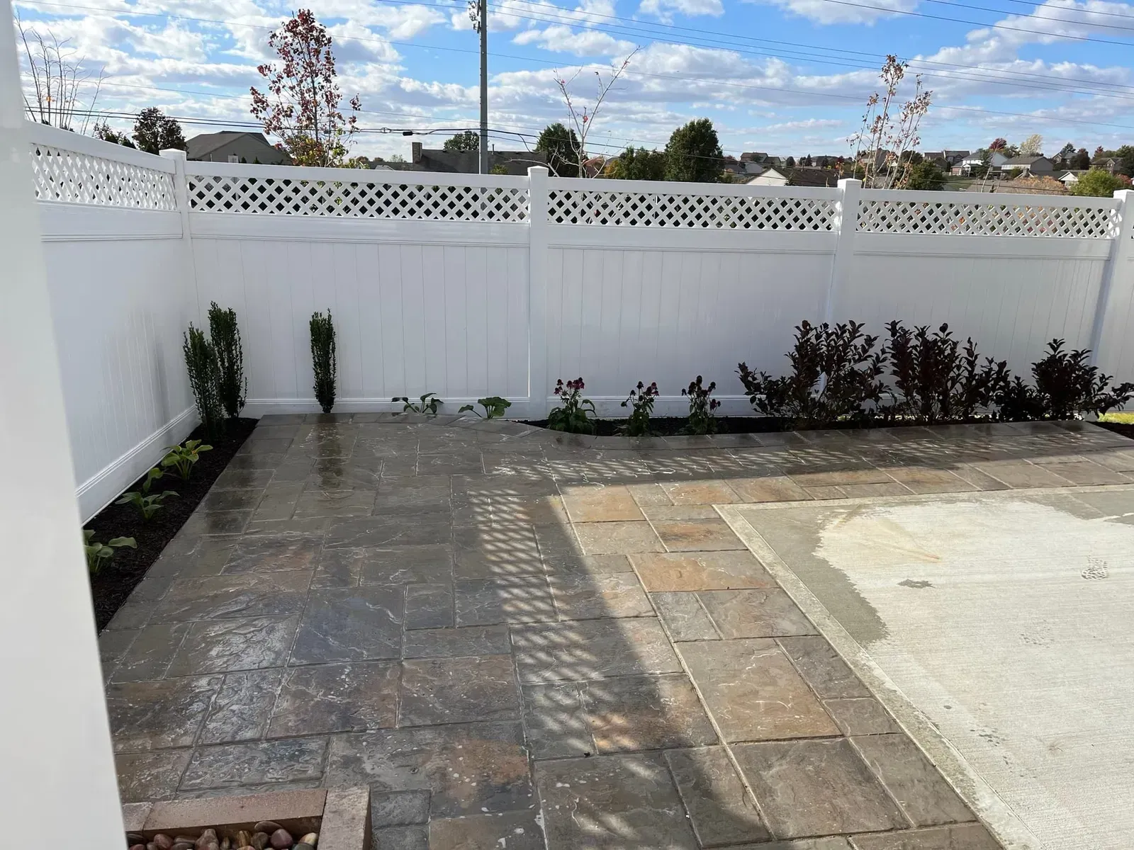 Small backyard with white lattice fence, brick patio, and newly planted shrubs under a sunny sky.